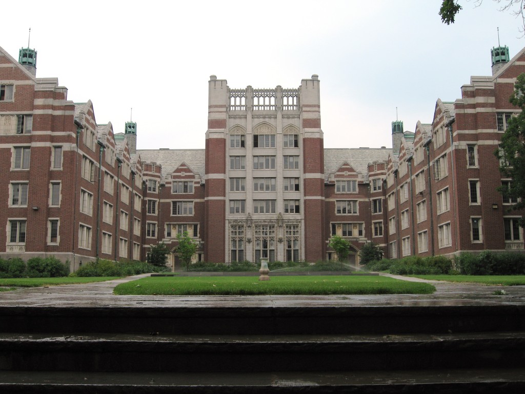 Symmetrical brick and stone university courtyard with a central green lawn, flanked by matching residential buildings with green-topped towers and a tall ornate central structure, viewed across a dark waterway.